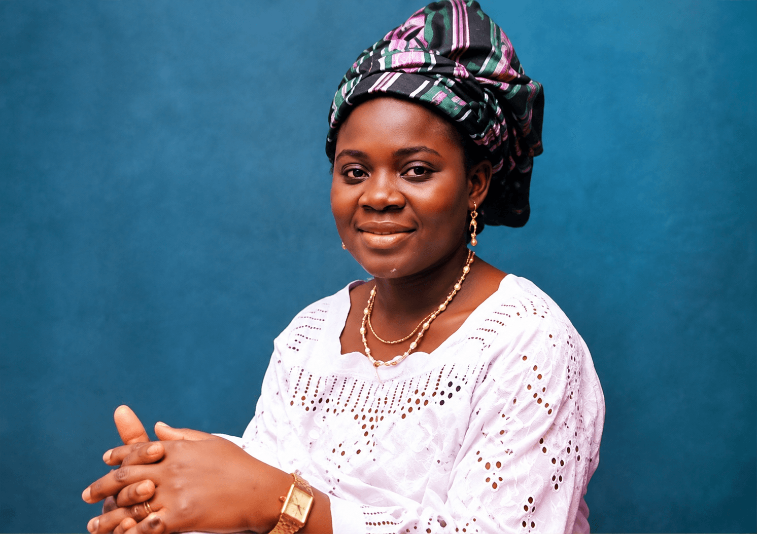 Studio portrait of Queen Salawa Abeni smiling in a striped gele and white eyelet buba, hands clasped, against a blue backdrop